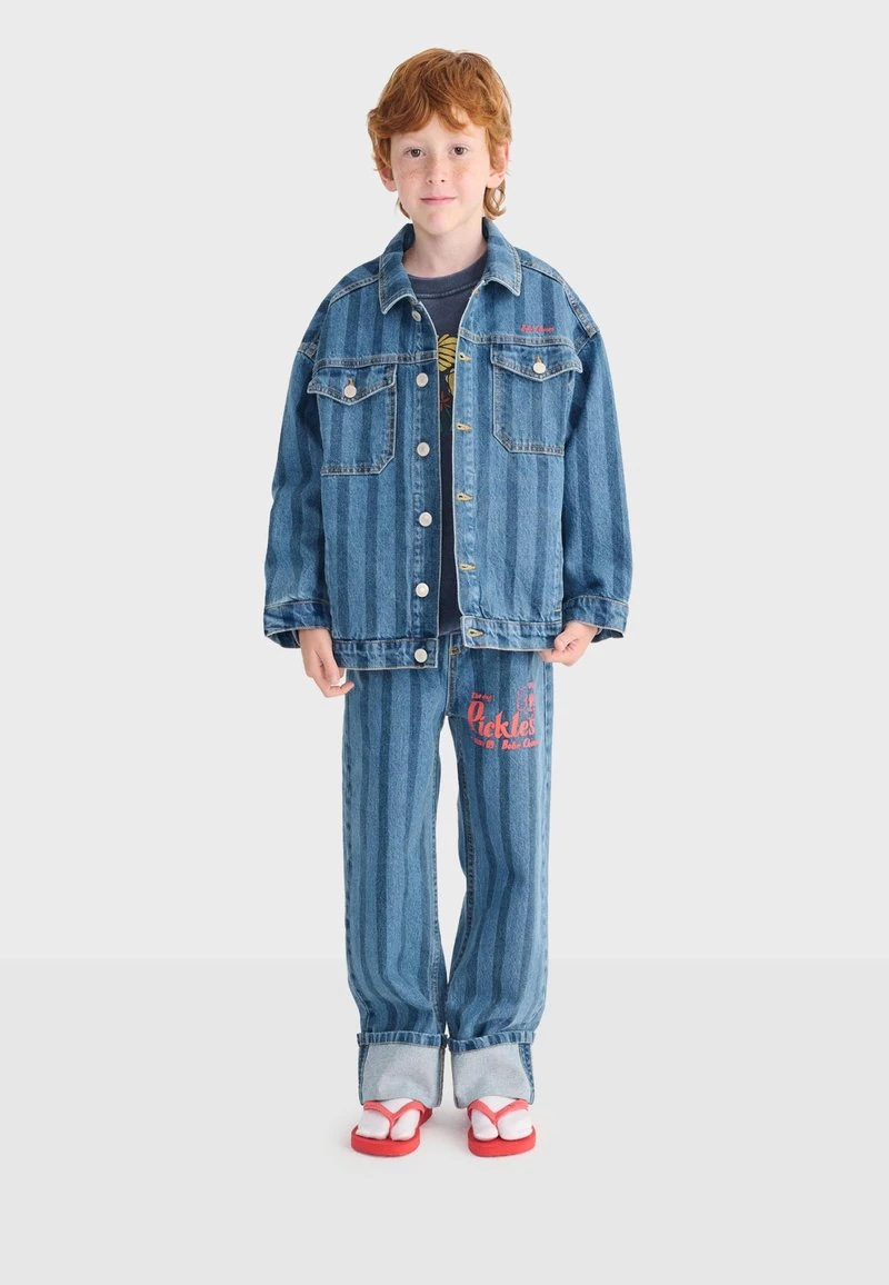 Roodharig kind met een blauw gestreept spijkerjasje en broek met rode tekst, zwart shirt en rood-witte sandalen, staand tegen een witte achtergrond.