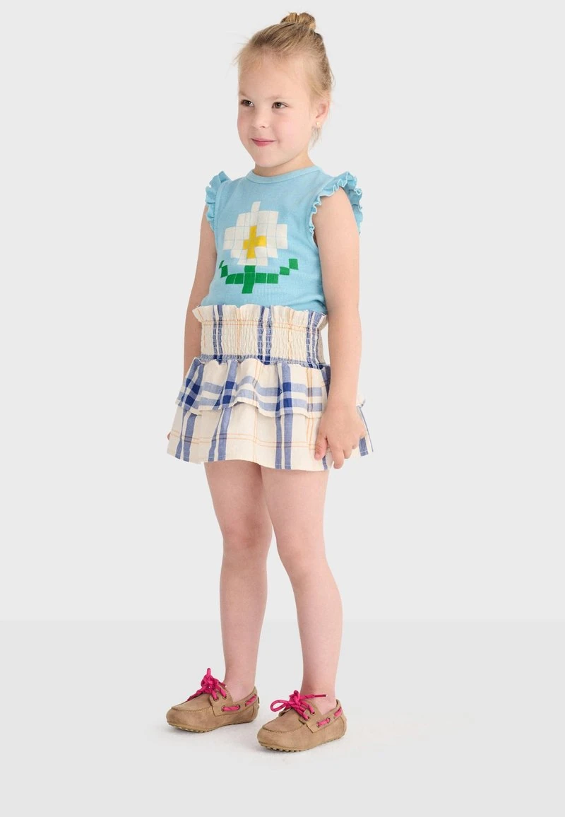Jong meisje met blond haar, gekleed in een blauw mouwloos topje met een witte bloem, een geruite rok met ruches en bruine schoenen met roze veters, staand en glimlachend.