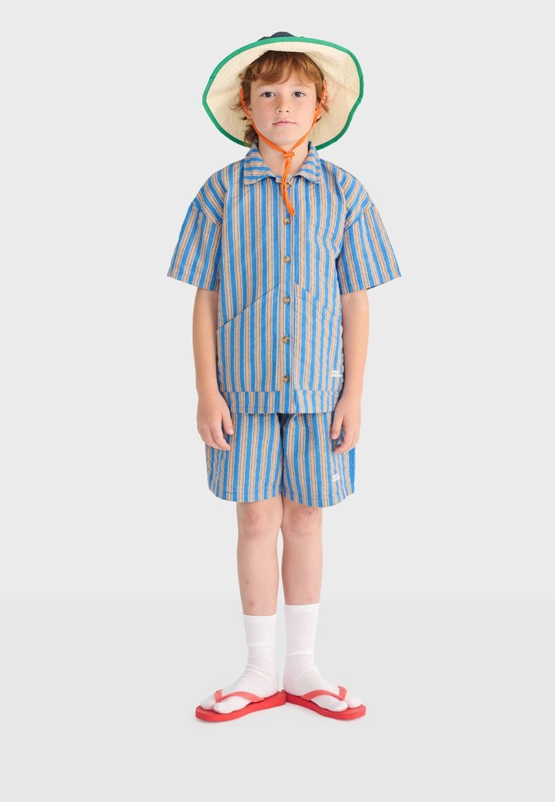 Kind draagt een gestreept blauw en oranje shirt met korte mouwen en korte broek, een zonnehoed met kinbandje, witte sokken en rode slippers, staat stil.