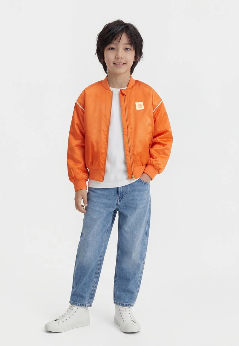 Kind met zwart haar, gekleed in een oranje bomberjack, wit T-shirt, blauwe spijkerbroek en witte sneakers, staand tegen een witte achtergrond.