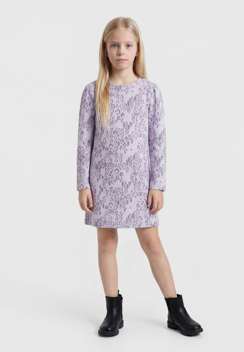 Jong meisje met lang blond haar in een lavendelkleurige lange mouw jurk met luipaardprint en zwarte enkellaarsjes, staand op een witte achtergrond.