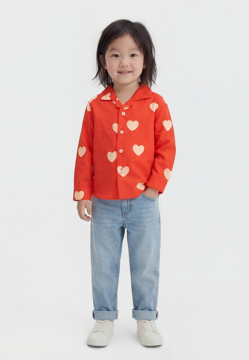 Jong kind met zwart haar dat een oranje shirt met lange mouwen en hartjespatroon draagt, lichtblauwe jeans en witte sportschoenen, glimlachend.