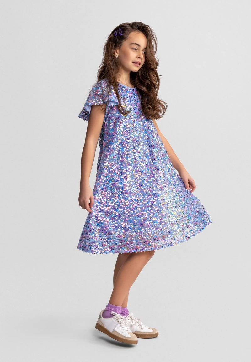 Jong meisje met lang haar, gekleed in een glanzende multicolor paillettenjurk, paarse sokken, witte sneakers en paarse haarclipjes, staand opzij.