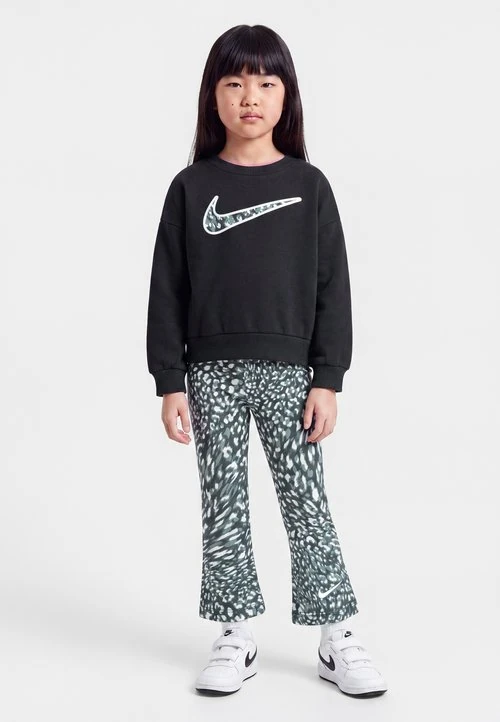 Jong meisje met stijl zwart haar, draagt een zwarte Nike trui, legging met patroon en witte Nike sneakers, staand tegen een effen achtergrond.