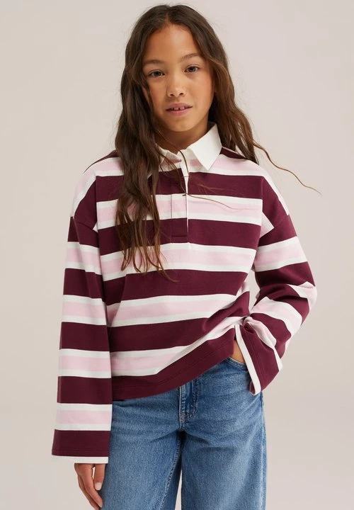 Jong meisje met lang bruin haar draagt een bordeauxrood, roze en wit gestreept shirt met lange mouwen en blauwe spijkerbroek, staand met één hand in de zak.