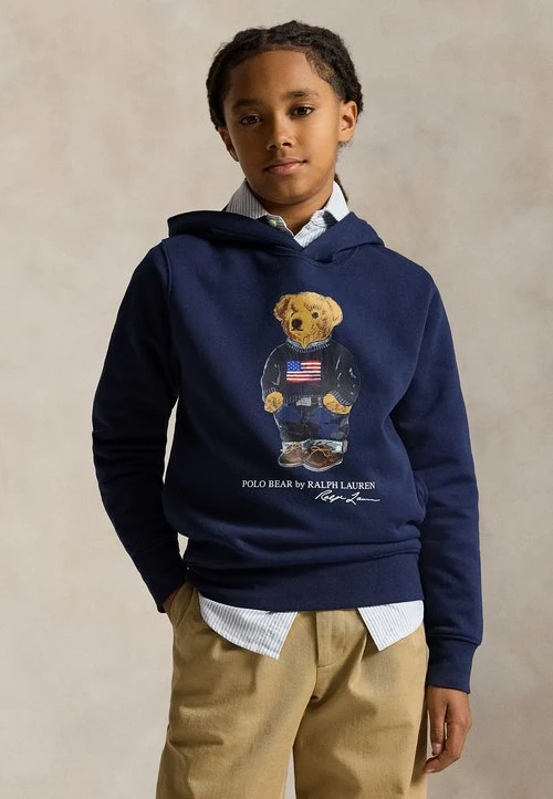 Kind met gevlochten haar dat een marineblauwe hoodie draagt met een teddybeer met Amerikaanse vlag, laag over een gestreept hemd en beige broek.